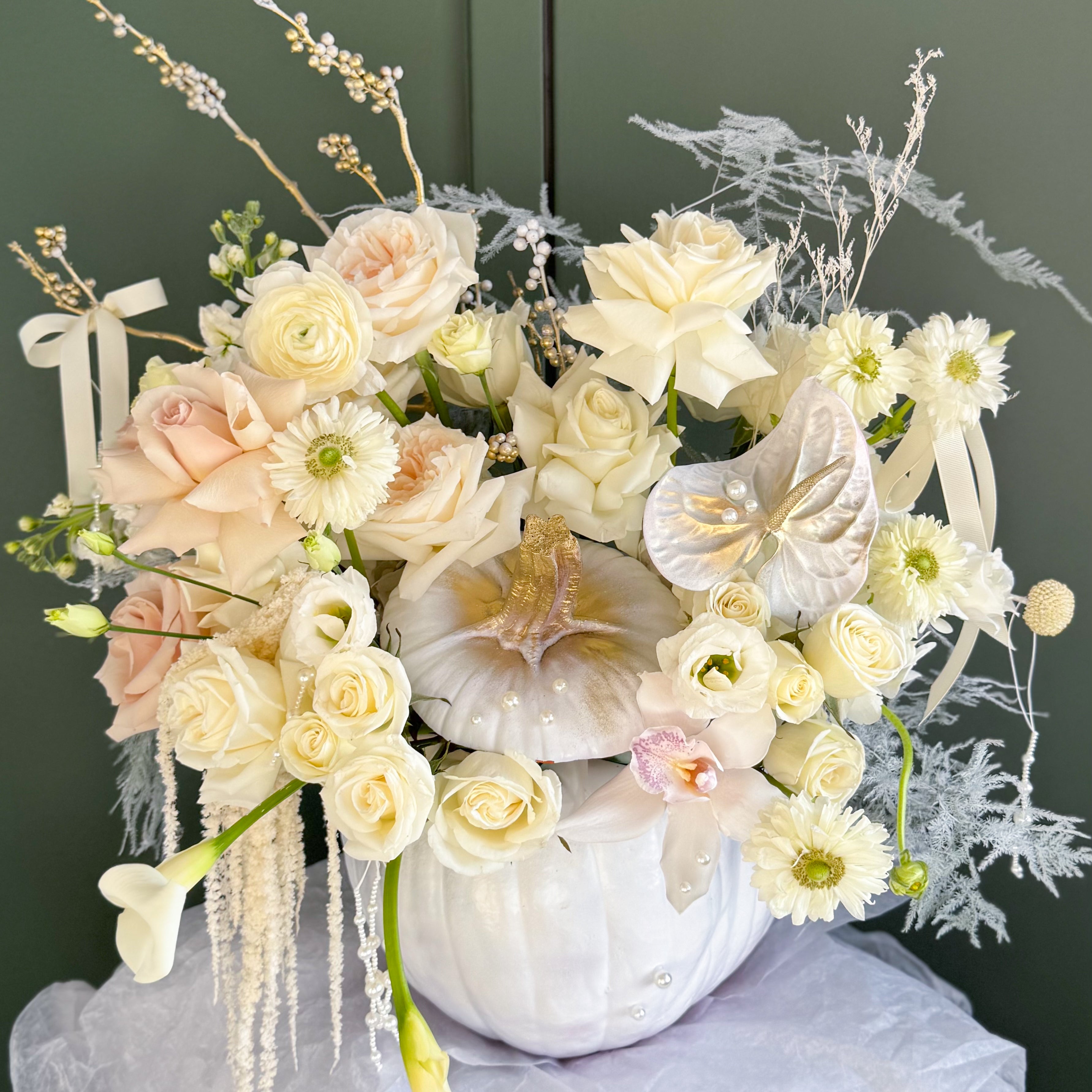 Luxury ivory and champagne floral arrangement featuring roses, orchids, and seasonal blooms in a decorative white pumpkin with pearl and gold accents.